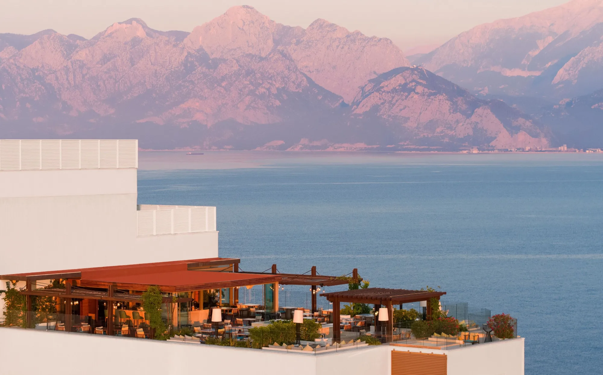 Terrasse eines Hotels mit Sitzplätzen, im Hintergrund das Meer und eine Bergkette während des Sonnenuntergangs.
