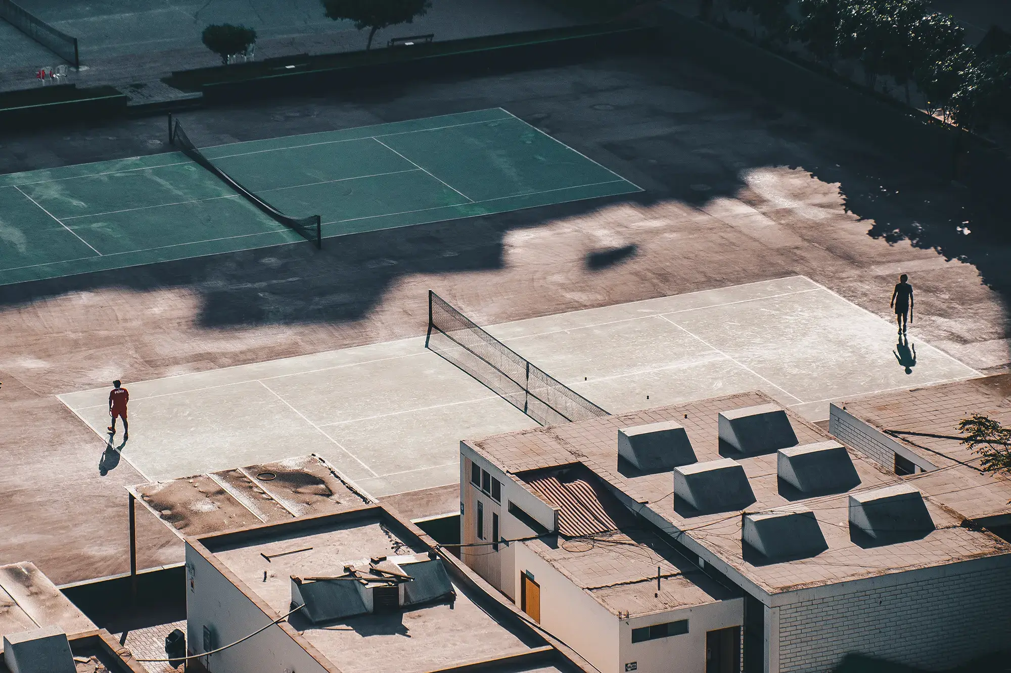 Zwei Personen spielen Tennis auf einem Platz in einem urbanen Umfeld mit Gebäuden im Vordergrund.