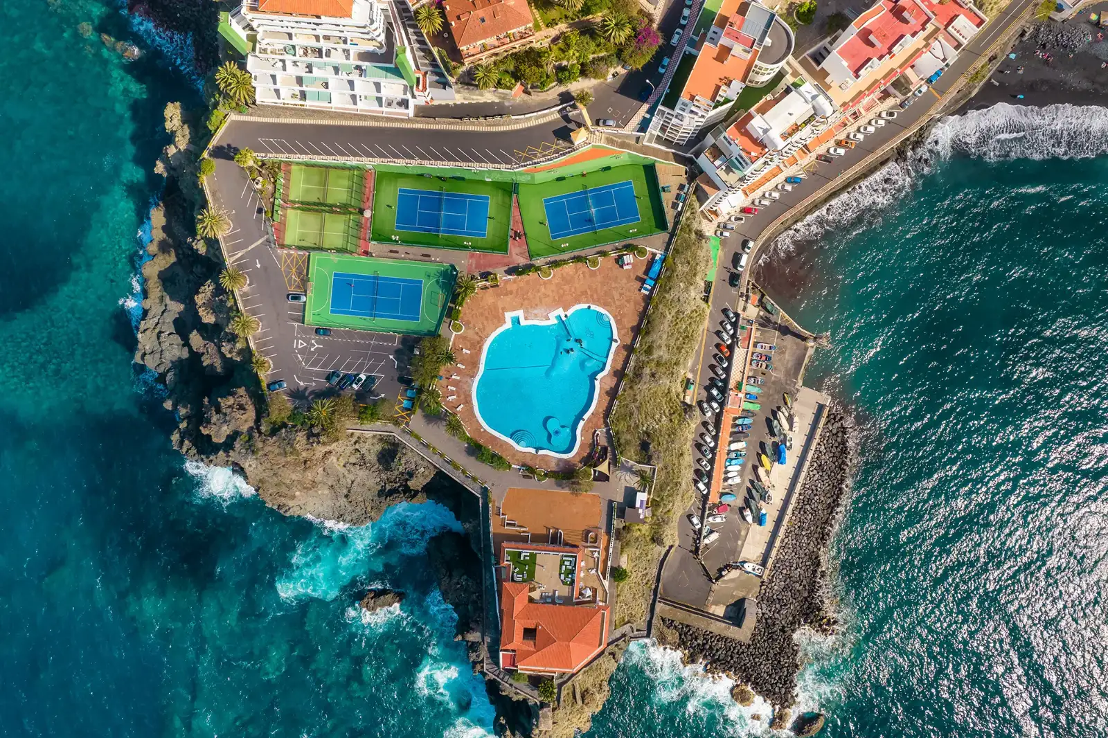Luftaufnahme eines Hotelkomplexes mit mehreren Tennisplätzen und einem Swimmingpool direkt an der felsigen Küste mit Blick auf das Meer.