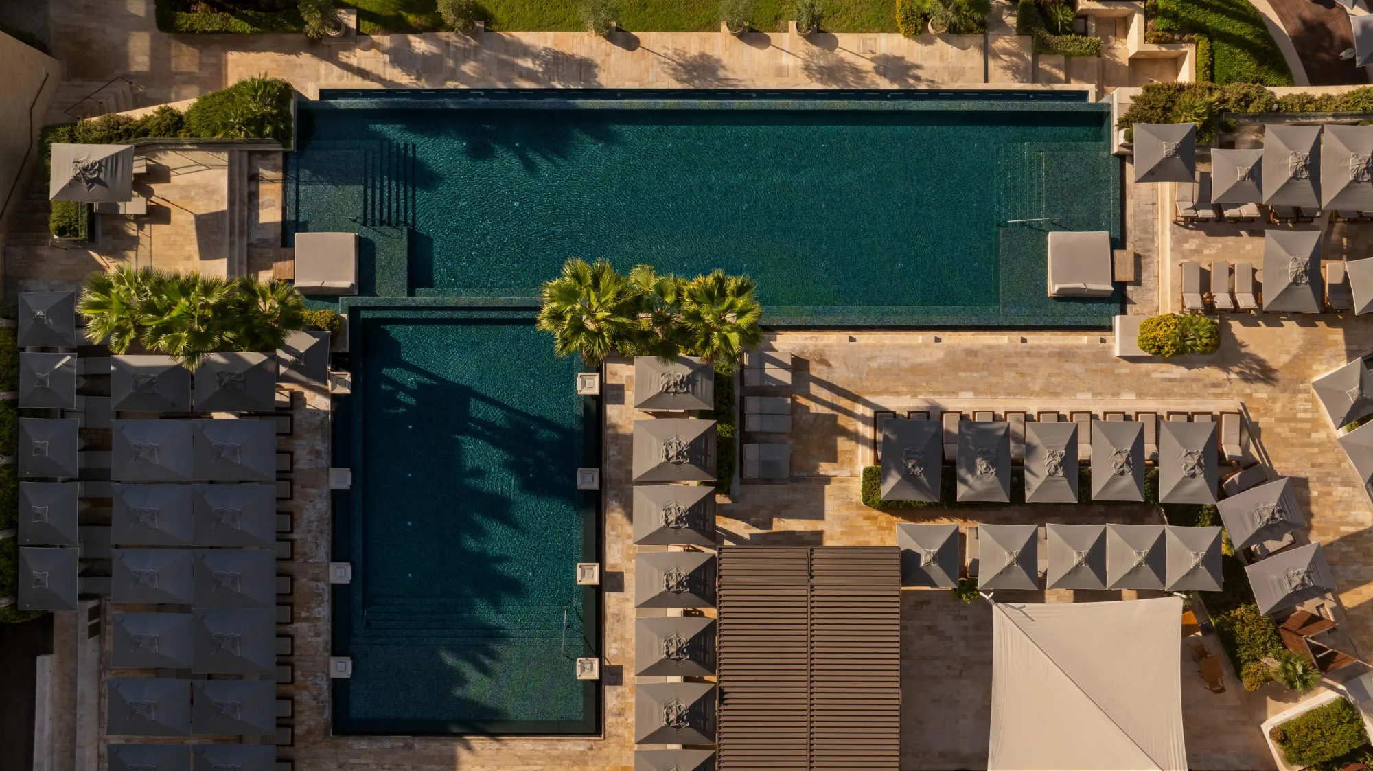 Luftaufnahme eines terrassenförmigen Pools mit Sonnenschirmen und Palmen in einem Hotelresort.
