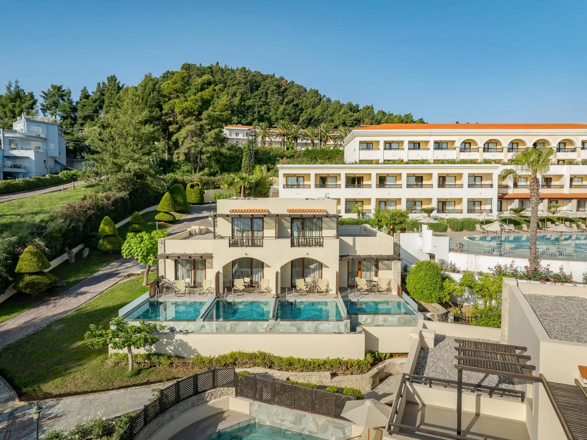 Blick auf ein Hotelgelände mit Zimmern, die private Pools und Terrassen haben, umgeben von grünen Bäumen und sanften Hügeln.