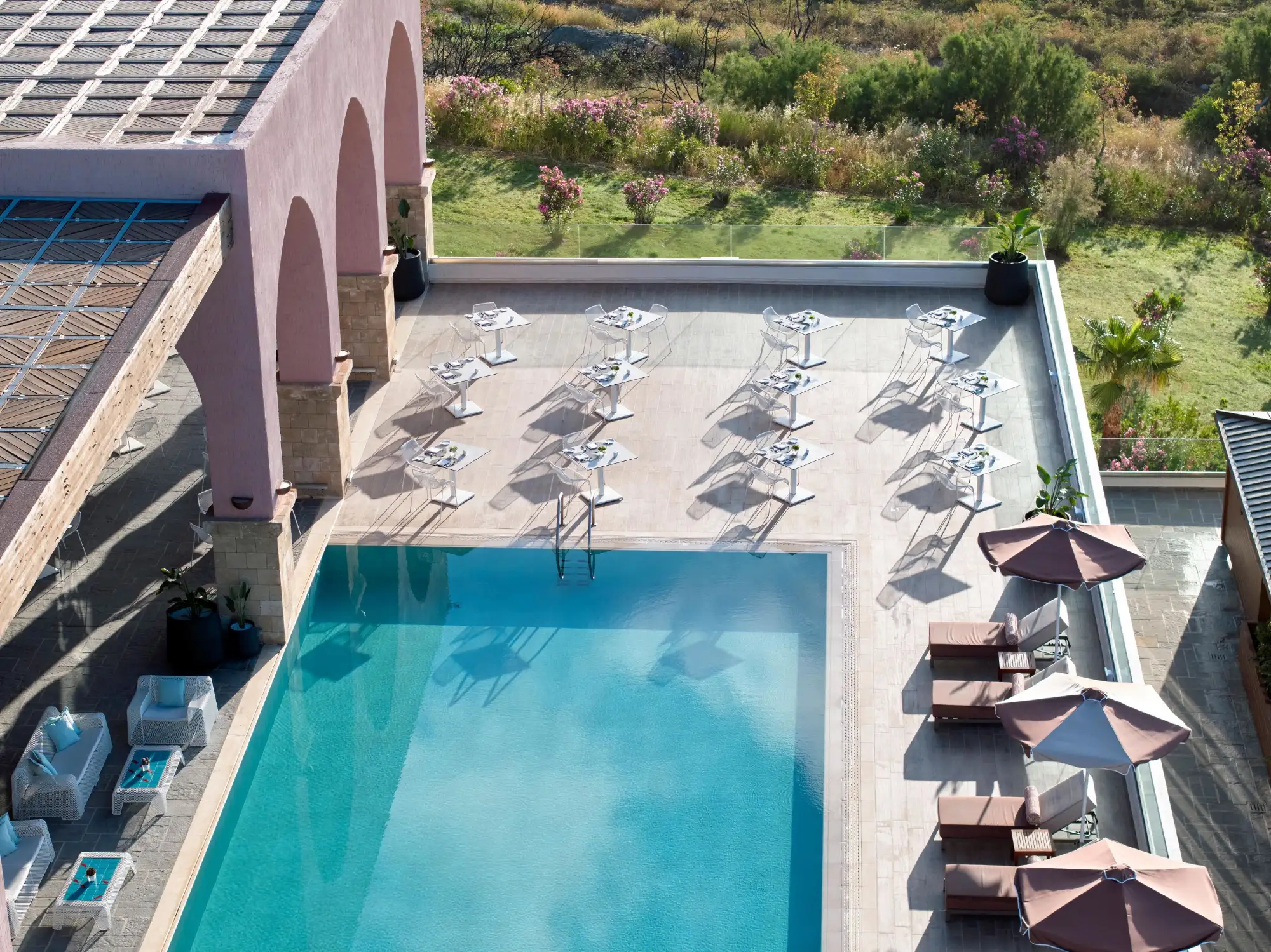 Blick von oben auf einen Hotelpool mit Liegestühlen und Schirmen neben einer Terrasse und Gärten im Hintergrund.