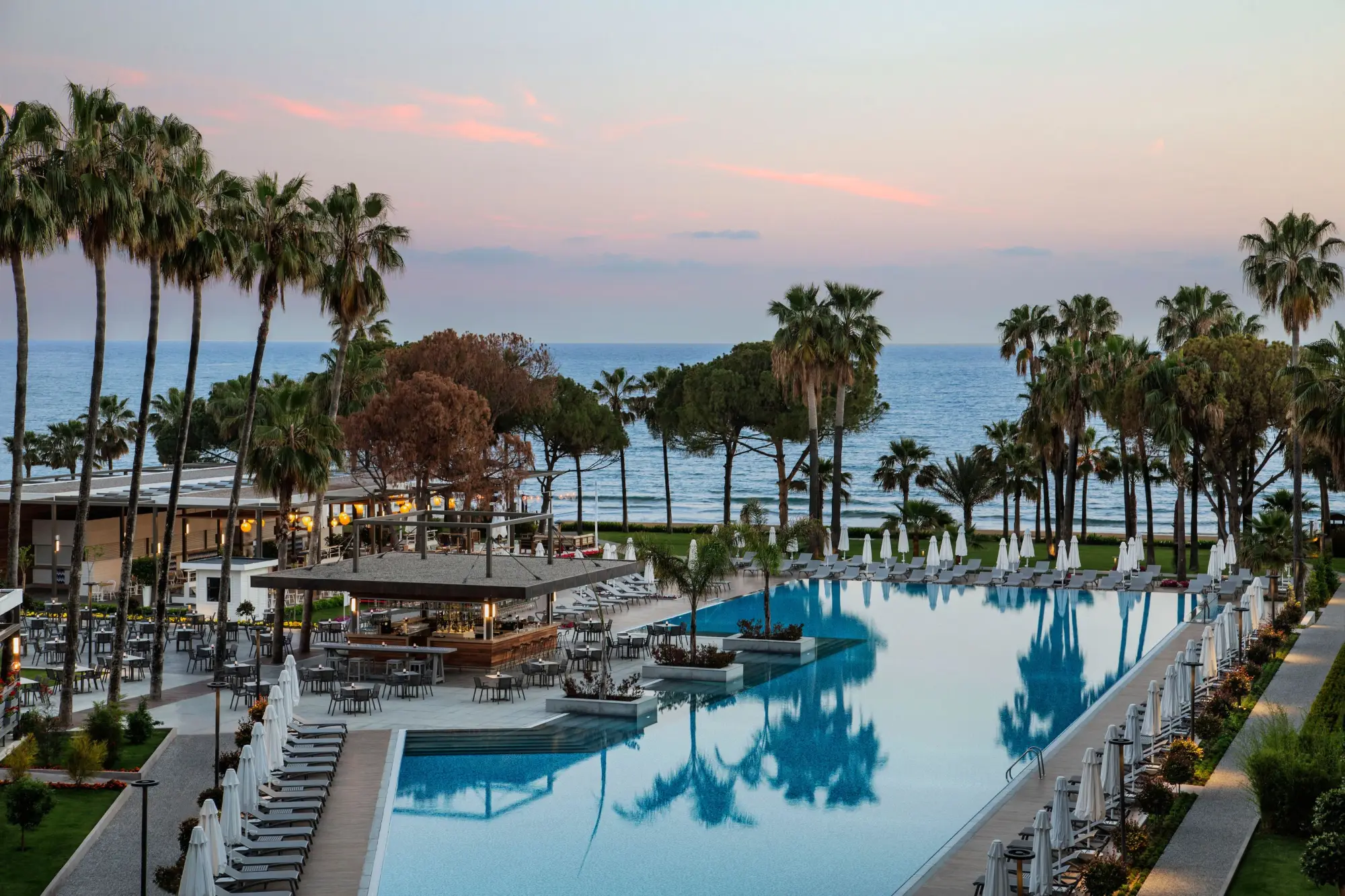 Großer Pool mit Liegen und Palmen am Meer bei Sonnenuntergang im Acanthus Cennet Barut Collection Hotel in Manavgat.